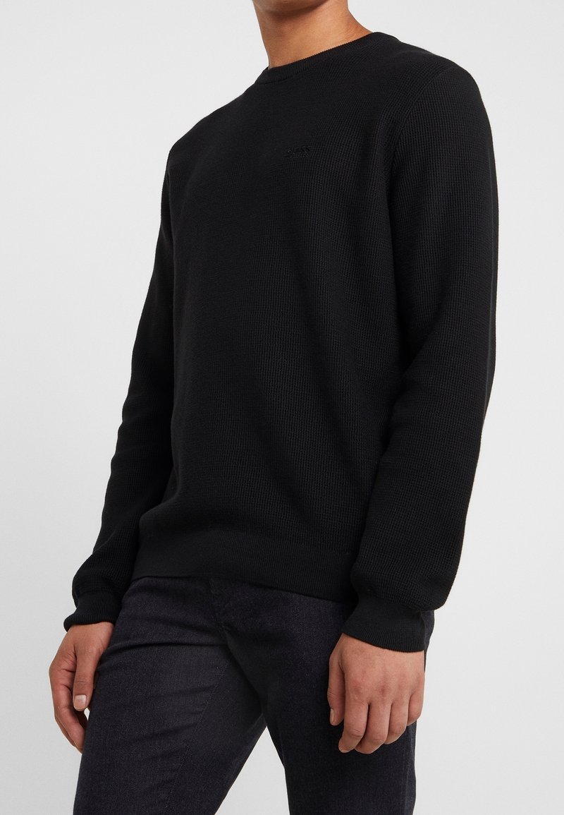 Black, ribbed knit sweater with a crew neckline and long sleeves, featuring subtle logo detailing on the chest. Worn with dark jeans.