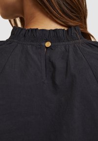 Blusa blu navy con scollo arricciato, dotata di una piccola chiusura con bottone rotondo dorato sul retro e un design plissè per un tocco di texture in più.