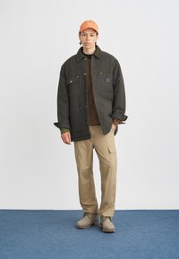 Chaqueta de cuadros negra de gran tamaño con collar verde, camisa marrón, pantalones de cargo beige y gorra de béisbol naranja. Combinada con botas de color claro.