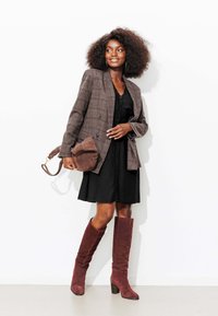 Blazer à carreaux marron sur une robe noire, accompagné de bottes en suède bordeaux montant jusqu'aux genoux et d'un sac à main en suède marron. Accessoires minimalistes, sur un fond blanc.