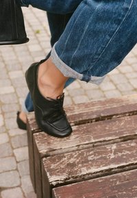 Personne portant un jean bleu retroussé et des chaussures en cuir noir posant le pied sur un banc en bois usé sur une surface pavée.