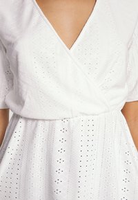 Femme portant une robe blanche en tissu œillet avec un corsage cache-cœur à col en V et des manches courtes, montrant la texture et le motif détaillés.