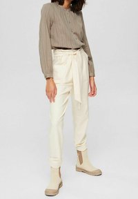Hellebeige Hose mit gebundenem Bund und gefaltetem Vorderteil, kombiniert mit einem strukturierten, gerippten, olivgrünen Langarmshirt. Beigefarbene Stiefeletten.