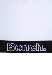 Haut de sport blanc avec une texture lisse, présentant une bande élastique noire en bas. La bande comporte le logo "Bench." en lettres blanches.