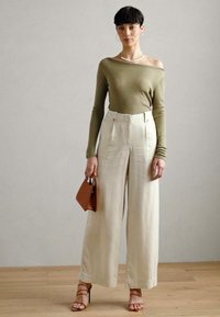 Olivengrønn off-shoulder topp, kremfargede vide bukser med frontfold, brun håndveske, og beige hæler med stropper på tregulv.