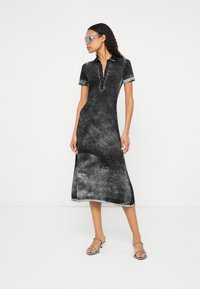 Femeie care poartă o rochie midi cu mâneci scurte, neagră și gri, sandale cu toc înalt argintii și ochelari de soare mari și transparenți, pozând pe un fundal alb.