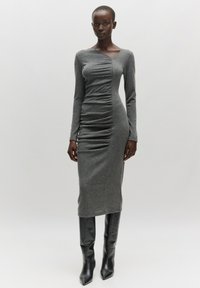 Graues Langarmkleid mit gerafften Details an der Vorderseite, figurbetonter Silhouette und glatter Textur, kombiniert mit schwarzen High-Heel-Stiefeln.