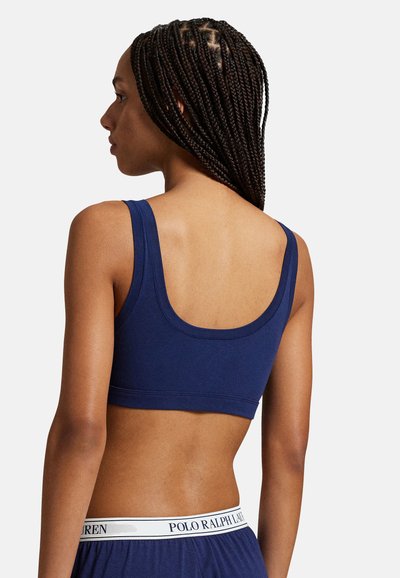Námořnicky modrý bralette z měkké látky se širokými ramenními popruhy a zaoblenými zády. Má kontrastní bílý pás s logem v oblasti pasu.
