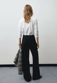 Un pantalon large noir associé à une chemise blanche à manches longues. La personne tient une veste gris à carreaux et a les cheveux bruns ondulés.