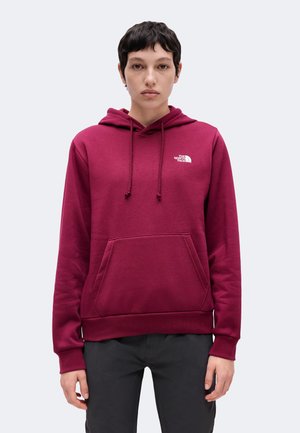 Personne aux cheveux courts et foncés portant un sweat à capuche bordeaux avec une poche avant et le logo The North Face sur la poitrine.