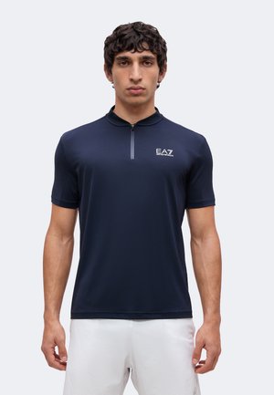 Homme portant un polo bleu marine à manches courtes zippé avec un logo blanc sur la poitrine et un pantalon blanc, debout devant un fond uni.