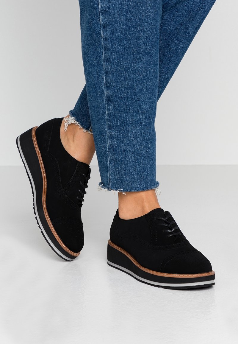 Chaussures en daim noires avec un bout arrondi, présentant des perforations décoratives et une semelle extérieure contrastante couleur tan. Portées avec un denim bleu cropped.