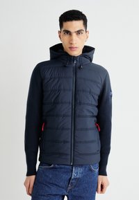 Granatowa kurtka puchowa z kapturem, pikowanej przedniej części, dzianinowymi rękawami, czerwonymi akcentami zamka i dopasowanym krojem. Posiada pionowe przeszycia i boczne kieszenie.