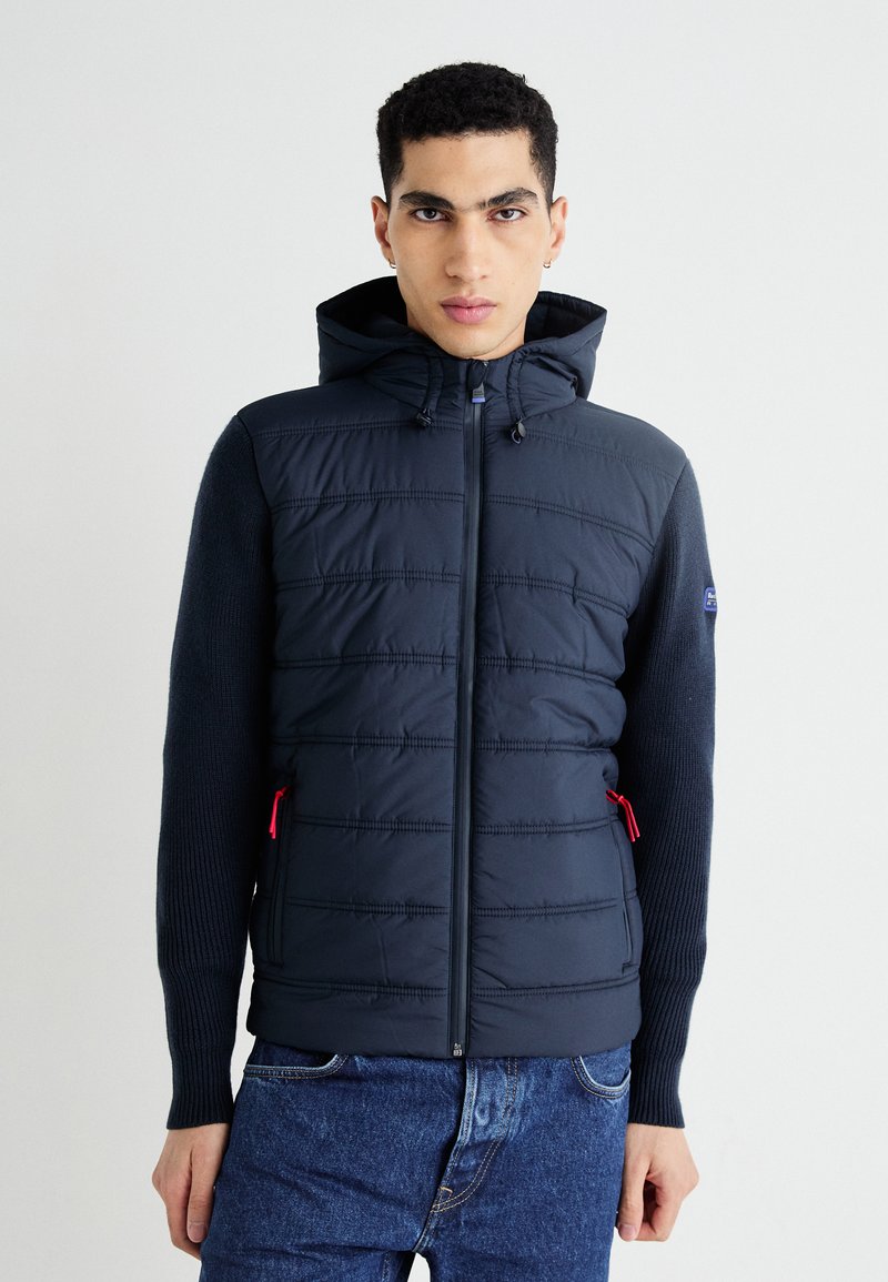 Granatowa kurtka puchowa z kapturem, pikowanej przedniej części, dzianinowymi rękawami, czerwonymi akcentami zamka i dopasowanym krojem. Posiada pionowe przeszycia i boczne kieszenie.