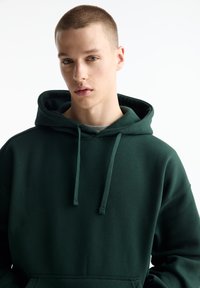 Dunkelgrüner Kapuzenpulli mit Kordelzug, aus weichem Material, mit einem schlichten Design und einer Kängurutasche.