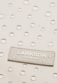 Druppels water verspreid over lichtbeige, gestructureerde stof met een rechthoekig label met verhoogde witte letters waarop "Larkson Hamburg" staat.