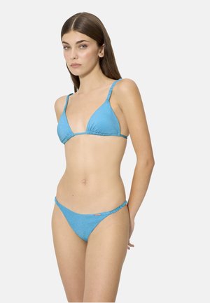 Mujer con cabello largo y castaño, vestida con un bikini azul claro texturizado con top de triángulo y braguitas de tiro bajo, posando frente a un fondo claro y liso.