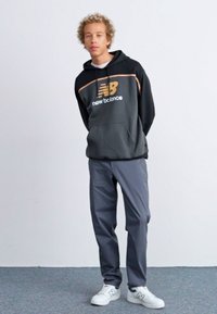 Sudadera negra con acentos grises y el logo de "New Balance", combinada con pantalones grises y zapatillas blancas, de pie sobre un fondo neutro.