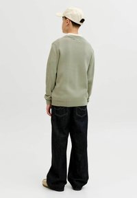 Jeune personne portant une casquette beige, un pull vert clair, un jean ample foncé et des chaussures beiges, debout de dos sur un fond uni.