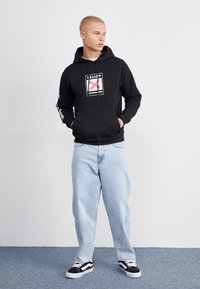 Sudadera negra con estampado gráfico, bolsillo tipo canguro y capucha con cordón, combinada con jeans rectos de color azul claro y zapatillas negras y blancas.
