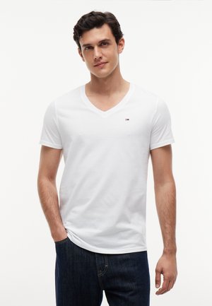 Ung mand iført en hvid V-hals T-shirt og mørkeblå jeans, stående med den ene hånd i lommen mod en ensfarvet hvid baggrund.