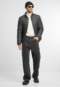 Sort pufferjakke med høj krave, cremefarvet turtleneck, mørke cargo-bukser og sorte støvler. Tilbehøret inkluderer en strikket hue og solbriller.