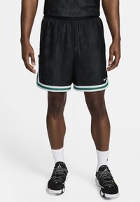 Atleta de pie con pantalones cortos de baloncesto negros con ribetes verdes y blancos, calcetines blancos y zapatillas deportivas negras.