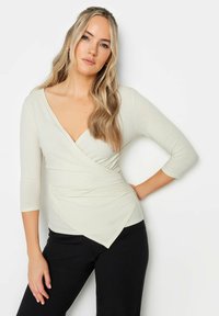 Cremefarbenes Wickeltop mit tiefem V-Ausschnitt, Dreiviertelärmeln, strukturiertem Stoff und asymmetrischem Saum, kombiniert mit schwarzen Hosen.
