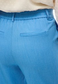 Modré texturované kalhoty se shromážděným elastickým pasem, obsahující dvě zadní kapsy a hladký povrch. Vhodné pro každodenní nošení.