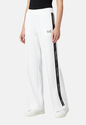 Pantalones blancos de pierna ancha con franjas laterales negras y logo EA7 Emporio Armani, usados con zapatillas blancas y una camiseta sin mangas negra.