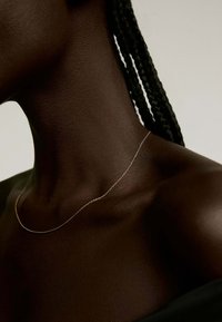 Collar delgado y delicado con un sutil diseño de cadena, con un acabado metálico elegante sobre piel oscura. Cabello trenzado parcialmente visible.