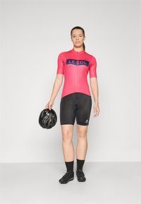 Sykkeltrøye i lys rosa med marineblå bokstaver, svarte shorts og hjelm. Tettsittende design med korte ermer og glatt tekstur.