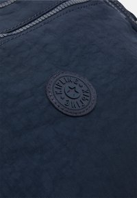 Navy blue fabric bag with a textured surface, featuring a circular black rubber logo patch embossed with "Kipling" and a star design.