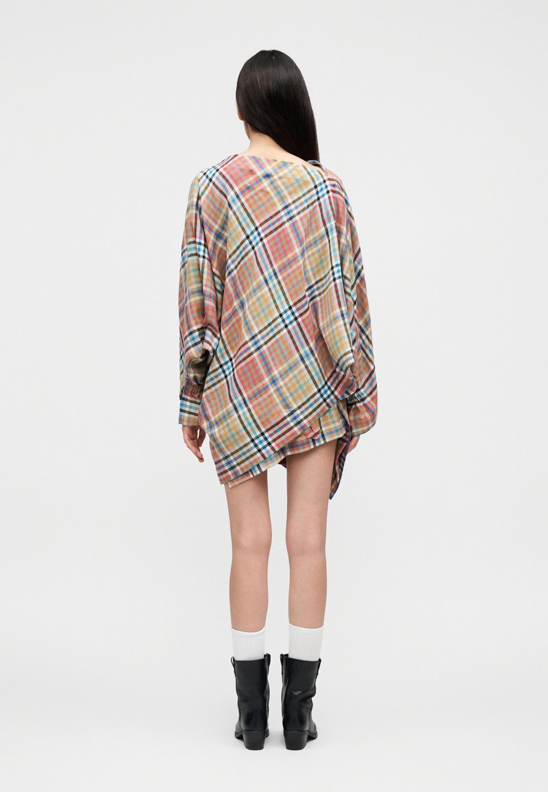 Femme debout de dos, portant une robe-chemise à carreaux colorés oversized, des chaussettes blanches et des bottines noires sur un fond uni.