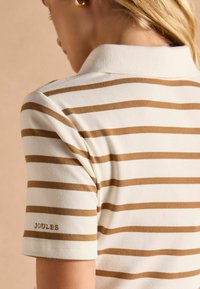 Pruhovaná polokošeľa v krémovej farbe s hnedými pruhmi, s rebrovanou textúrou a preloženým golierom. Na rukáve je vyšité logo "JOULES".