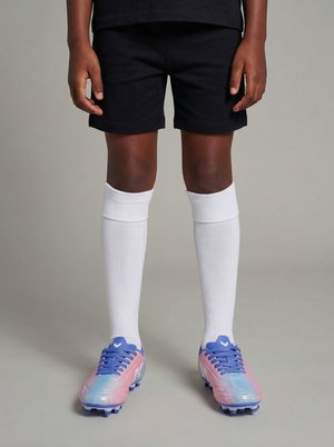 Barn står iført sorte shorts, lange hvide sportssokker og pink og blå fodboldstøvler på en ensfarvet grå baggrund.