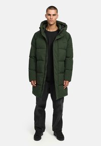 Giacca puffer verde oliva con cappuccio, caratterizzata da un design trapuntato, chiusura frontale con zip e forma leggermente allungata. Indossata sopra una maglietta nera.