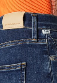 Detalle trasero de unos jeans de denim con un parche de cuero marrón que dice "CITIZENS of HUMANITY," una pequeña etiqueta de tela blanca que dice "C of H," y una parte superior de canalé naranja.