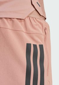 Shorts de sport roses en tissu texturé, avec une poche zippée et des accents rayés noirs sur un côté.