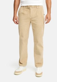 Beige corduroy pants with a straight cut, featuring front pleats and side pockets. Worn with white sneakers and a fitted white shirt.