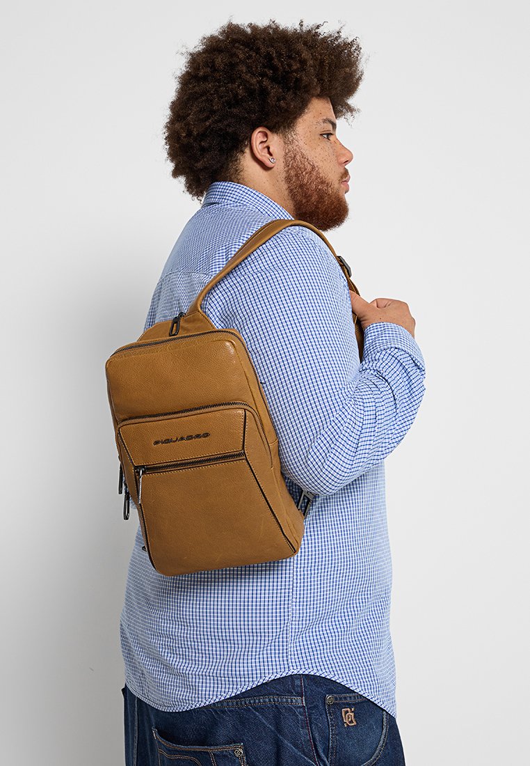 Uomo con capelli ricci e barba, che indossa una camicia a quadri blu e jeans, mentre porta a tracolla uno zaino a tracolla in pelle color beige.