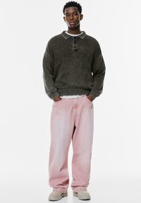 Donkergrijze fleece trui met een kraag, knoopdetails en een oversized pasvorm. Gecombineerd met lichtroze baggy jeans en beige schoenen.