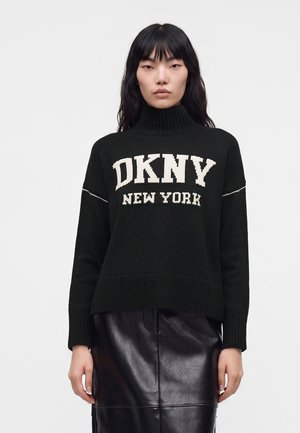 Vrouw draagt een zwarte coltrui met de tekst "DKNY New York" en een zwarte leren rok tegen een effen witte achtergrond.