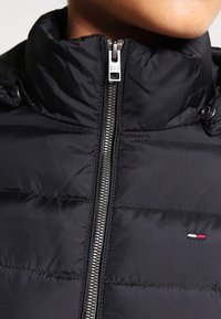 Czarna kurtka puchowa z wysokim kołnierzem, z szarym zamkiem błyskawicznym i małym logo w trzech kolorach. Gładka, wodoodporna faktura materiału.