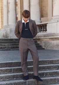 Pruun ruuduline ülikond musta polosärgiga ja valge kraega, selle juurde sobitatud tumedad kingad, seistes välitingimustes kivist trepil.