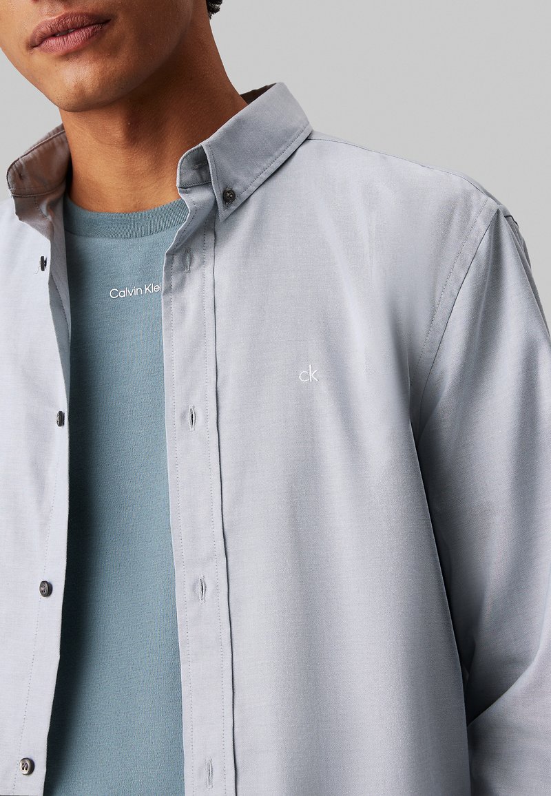 Camisa de botón de color gris claro con cuello, que presenta pequeños botones negros y un sutil logo "ck". Llevada sobre una camisa azul.