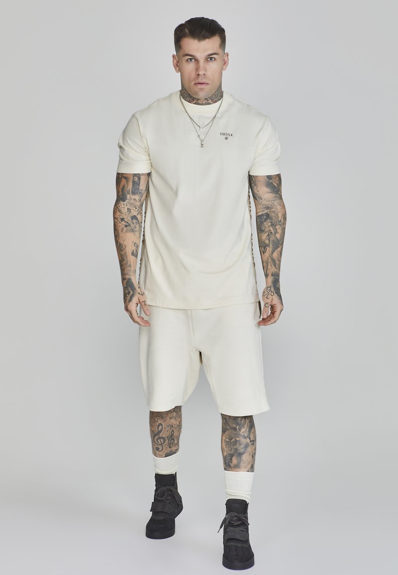 Camicia a maniche corte color crema e pantaloni corti realizzati in tessuto morbido. Presenta un piccolo logo sul petto e una vestibilità comoda. Indossati con sneakers nere.