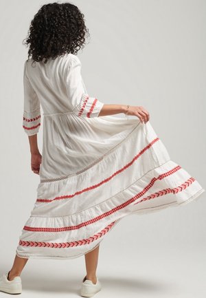 Largo vestido blanco con mangas tres cuartos, adornado con franjas rojas bordadas y un diseño de volantes en capas. Estilizado con zapatillas blancas.