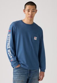 Blaues langärmeliges Baumwoll-T-Shirt mit Brusttasche, weißem Aufdruck 'WESTERN WEAR' am Ärmel und einem kleinen Logo-Patch auf der Tasche.