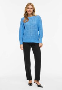 Femme portant un pull en tricot bleu vif avec un motif texturé, un pantalon noir à jambes droites et des talons noirs à bout pointu, debout devant un fond blanc.